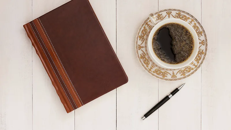 Braunes Notizbuch, Kugelschreiber und eine Tasse Kaffee auf einem hellen Holztisch